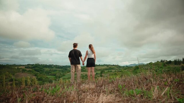 Romantic young couple holding hands enjoying the breathtaking panoramic view of the tuscan hills in italy and contemplating the future together on a cloudy day
