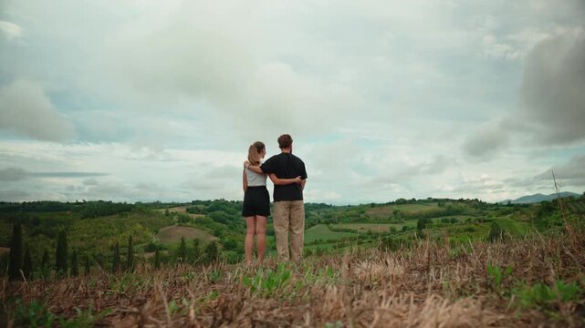 Young couple hugging and enjoying the scenic view of the beautiful rolling hills of tuscany, italy, while pointing at the horizon on a cloudy day from a hilltop