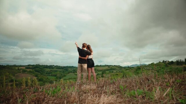Romantic couple embracing while enjoying the breathtaking view of the green hills of tuscany, italy. Man pointing at the idyllic landscape during a summer travel holiday