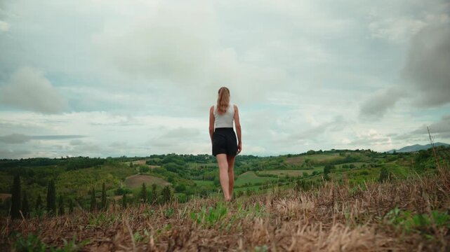 Woman walking barefoot toward a scenic view of the tuscan countryside, feeling freedom and joy as she raises her arms in celebration of nature's beauty and her escape