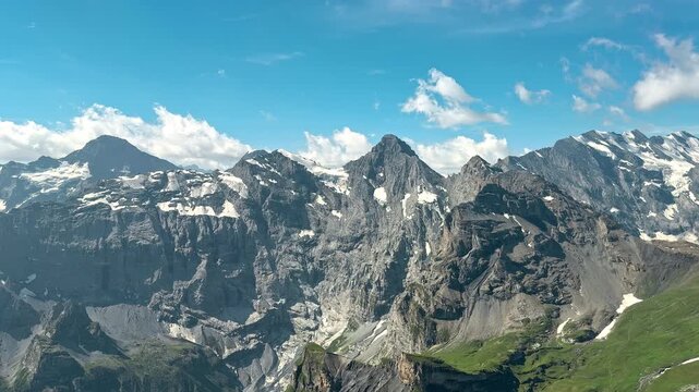 Rocky Swiss Alps rise beneath white clouds and patches of summer snow, Elevated travel footage surveys remote ridges and green mountain slopes