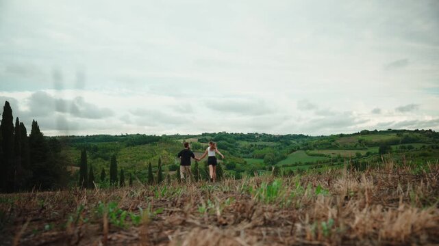 Young couple running hand in hand through a field enjoying a romantic holiday vacation in the beautiful green hills of the tuscan countryside in italy, a slow motion shot