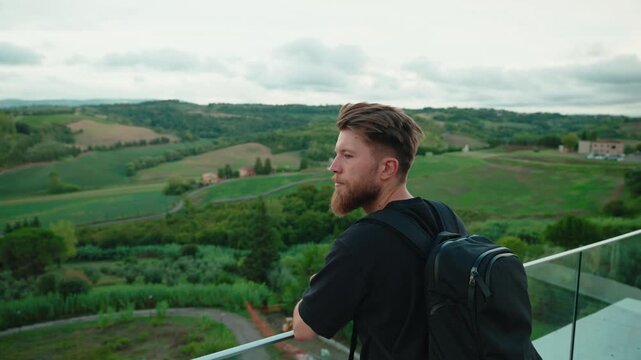 Young bearded traveler with a backpack enjoying the beautiful green rolling hills of tuscany from a scenic viewpoint during an adventurous italian holiday vacation