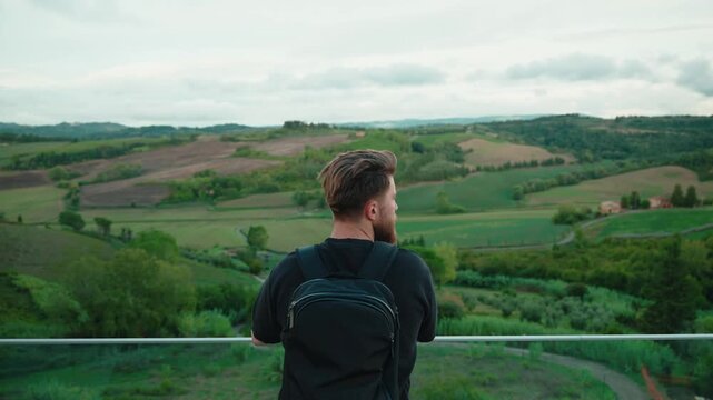 Young bearded traveler with a backpack enjoying the beautiful and peaceful green landscape of the tuscany hills, admiring the idyllic italian countryside scenery