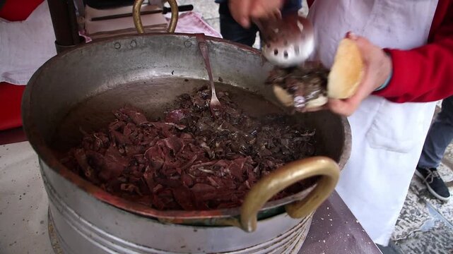 Close-up of a street vendor in Palermo, Sicily, filling a traditional bread roll with simmered spleen and offal from a large copper pot to make the local delicacy Pani ca Meusa.