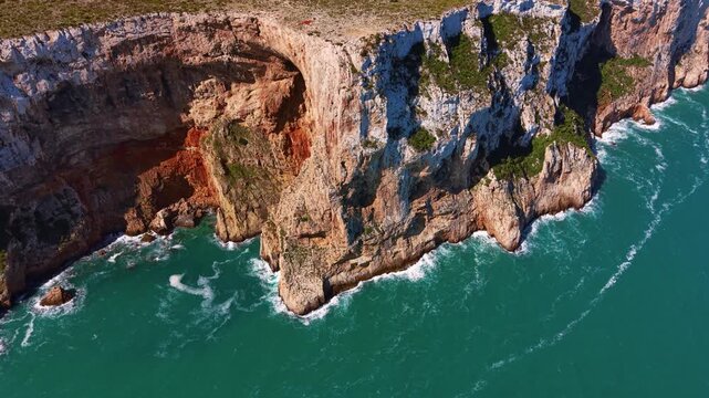 Dramatic coastal cliffs with layered rock formations plunge into vivid turquoise ocean waters, waves crashing at the base with patches of green vegetation on rugged cliff tops