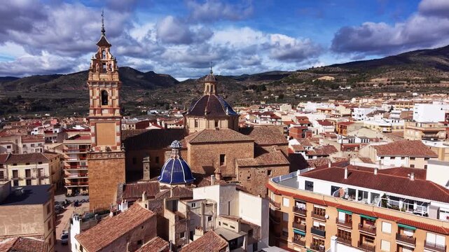Panoramic view of Chelva, Spain, featuring a historic church bell tower and tiled dome rooftops amid a hillside town under a dramatic cloudy sky
