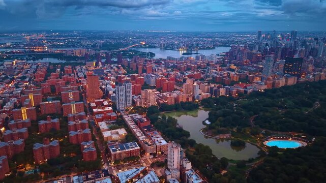 Part of Central Park with lush greenery and ponds surrounded by dense cityscape with red brick buildings. Heavy dark clouds gathered over the city in the sky.