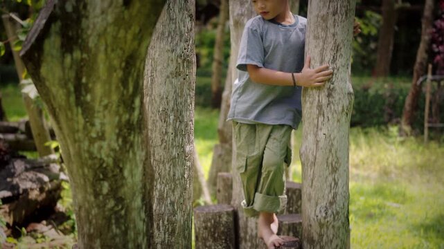 Young Asian child walking barefoot and balancing across a wooden log obstacle course in a sunny green park, developing coordination, balance, focus, strength, and motor skills through outdoor play.