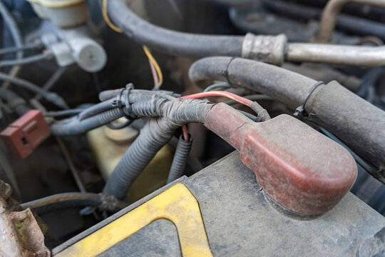 Close up of a car battery positive terminal with a protective red cover and electrical wiring harness in an engine bay, highlighting vehicle maintenance and repair.