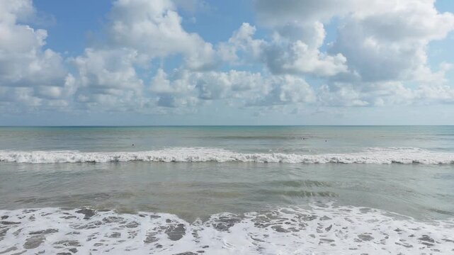 Aerial view of ocean waves rolling towards the shore under a bright blue sky with scattered clouds in Costa Rica.