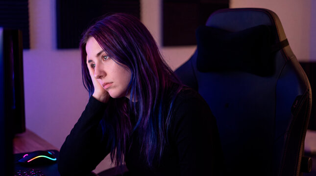 Tired and frustrated young female gamer with purple hair sitting in a dark neon-lit setup feeling bored or defeated after losing an online match.