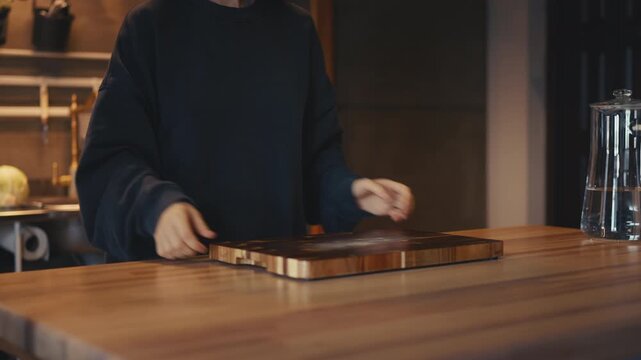 Close-up of a woman moving a cutting board, glass bowl, cabbage, and knife from the kitchen counter onto a wooden table for food preparation. Healthy dinner and cooking concept in a cozy, warm home.