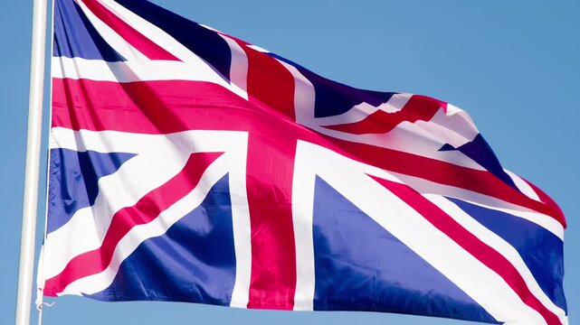 British Flag Waves the Wind. The Big State Flag is illuminated by the sun and flutters epically in the wind against the blue sky