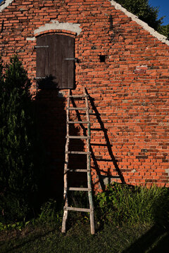 A moody, nostalgic, and mysterious view of a rural red brick wall. A weathered wooden ladder leads up to a closed, dark loft door, casting a deep, eerie shadow in the fading warmth of the sunset.