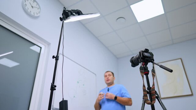 Modern camera on tripod and lights directed on the man in blue uniform. Low angle view at the doctor talking for video.