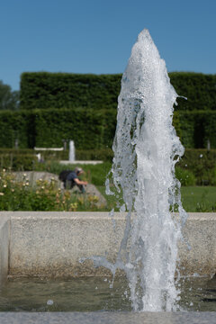 Water fountain sprays powerful vertical stream with drops in concrete basin. Clear liquid splashes inside public park during hot summer day. Urban architecture and landscape decoration.