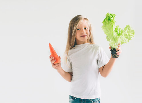 A girl in a white shirt isolated on a light background holding carrot and salad. Smiling child loves fruits and vegetables