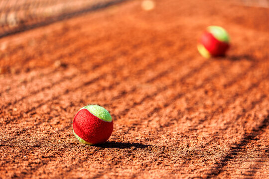 clay tennis court with scattered two-tone balls under bright sunlight. The net and long shadows create a dynamic atmosphere, emphasizing movement and competition