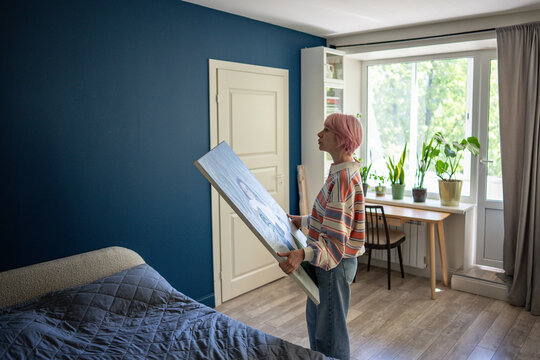 Pensive woman holding large canvas artwork in hands while standing in bedroom looking at wall, thoughtful female artist in apartment bedroom with painting in hands thinking and choosing place to hang.