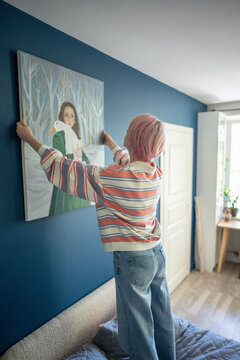 Focused woman artist standing on bed in bedroom and adjusting large canvas painting on wall, creative female painter hanging own created artwork in room, living space design and DIY interior.