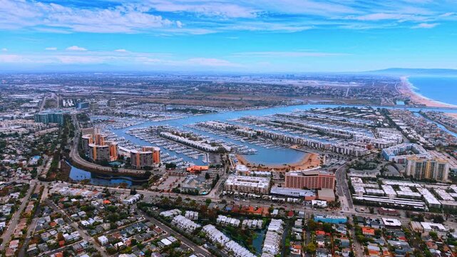 Marina with yacht berths and waterfront apartment buildings. Rows of boats fill a harbor surrounded by housing, with the Pacific coastline in the distance.