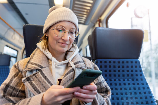 Portrait young adult woman passenger use smartphone inside modern train wear winter hat glasses coat commuting on sunny day. Casual travel lifestyle scene public transport interior urban routine