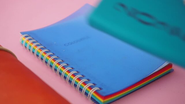 Close up hand picking up turquoise notebook from stack of colorful stationery items on pink background.