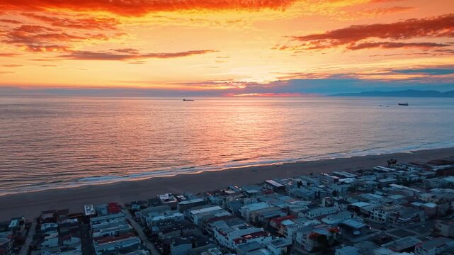 Sunset over the ocean and coastal homes in Los Angeles. Orange clouds reflect on the water above a beach and rows of buildings seen from the air.