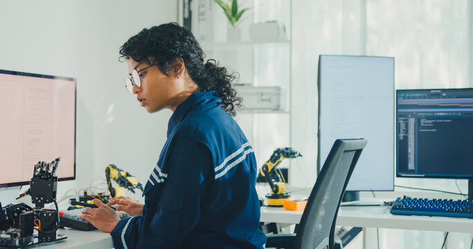 Young African American female robotics engineer coding AI software for robotic arm in laboratory. Embedded system, firmware testing, machine learning, industrial automation, future technology concept.