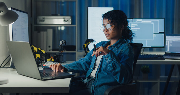 Young African American female programmer coding AI software on multiple screens in night office. Network security, cybersecurity, machine learning, working late, overtime, robotics automation concept.