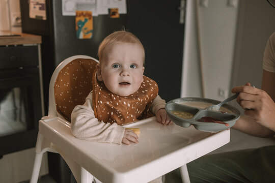A close-up shot of a parent's hand gently feeding a sweet infant child with a small spoon. The baby is sitting in a white highchair, wearing a patterned silicone bib, and curiously looking ahead