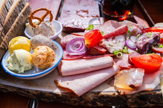 Close view of traditional Austrian Bavarian Brettljause alpine snack board served on a wooden platter, with cured meats, sausages, cheeses, tomato, cucumber, red onion and mini pretzel