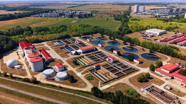 Aerial view of wastewater basins and circular clarifiers. Parallel treatment channels, white tanks and red roofs fill a site bordered by woodland.