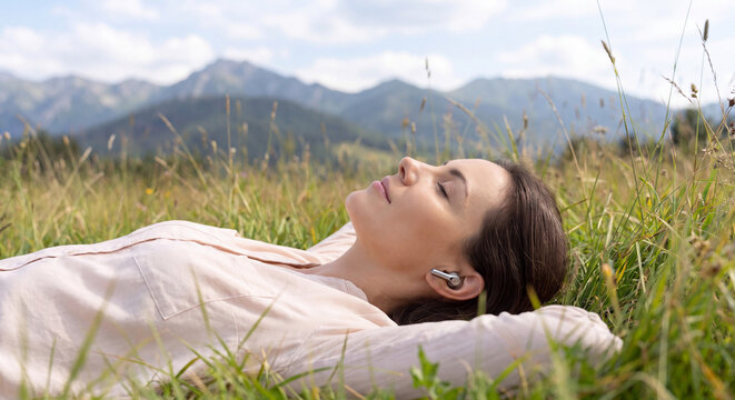 A serene woman rests lying down on a lush alpine meadow with eyes closed wearing headphones against a majestic panoramic mountain range under a blue sky