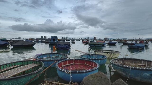 Composite timelapse of fast-moving clouds over traditional Vietnamese coracle boats gently swaying on the water at a fishing harbor in Da Nang, Vietnam, on an overcast day.
