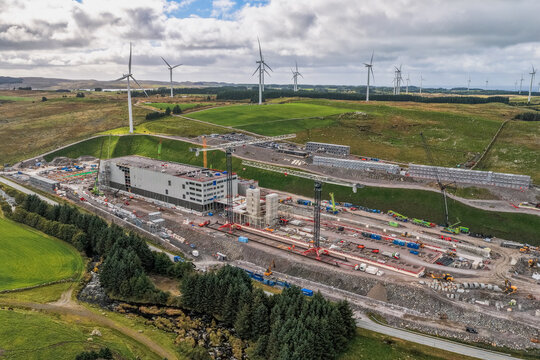 Aerial view of the Green Mountain data center construction site featuring large industrial buildings, tall cranes, and surrounding wind turbines on green hills in Undheim, Rogaland, Norway.
