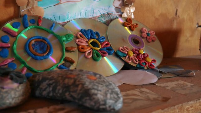 Handheld camera shows colorful kids crafts made from clay and CDs on a wooden surface in Khakassia, showcasing imagination and recycling art.