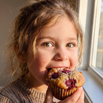 Close-up portrait of a joyful young child with crumbs on her face, delightedly eating a raspberry muffin in sunlight.