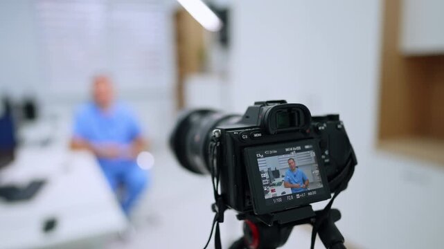 Rear camera screen framing a healthcare presenter. A man in blue scrubs appears on the display, with the seated subject and desk blurred in the background.