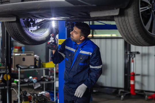 man professional auto mechanic inspecting raised car undercarriage with flashlight in repair garage, checking wheel, suspension, brake components for vehicle maintenance and safety service.