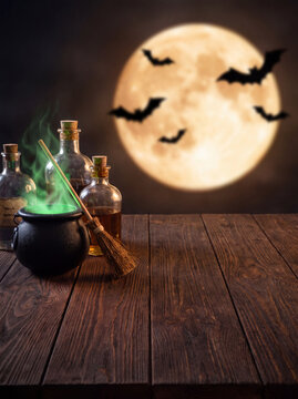 A rustic wooden table holding a bubbling green potion cauldron near glass bottles and a magic wand with a glowing full moon and bats in the dark night background