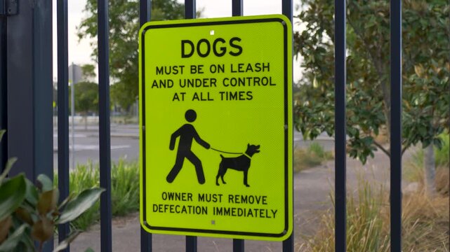 Slow motion close-up of yellow municipal dogs on leash and waste to be cleaned up immediately regulatory rules sign with character symbols mounted on metal gated fence at public oval park in Australia