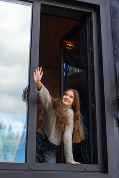 Young woman waving from an open window of a modern house, cheerful lifestyle portrait with long hair and casual clothes against reflective glass.