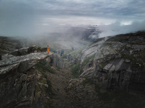 Aerial view of a person in a yellow coat standing on a rocky ledge overlooking the dramatic cliffs and misty depths of Hellmojuvet canyon in Hamaroy, Nordland, Norway.