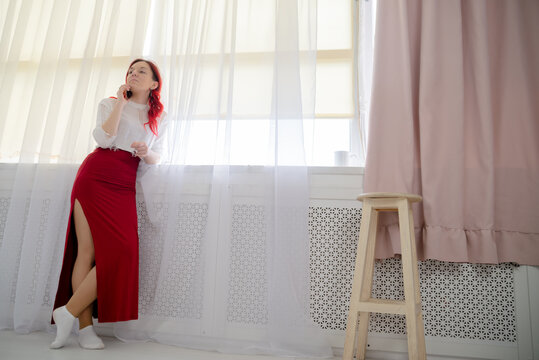 Young woman with red hair talking on smartphone by the window at home