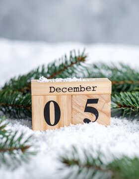 Wooden calendar cubes with text DECEMBER 5 on a snow-covered rustic surface, fresh fir tree branches, cozy festive atmosphere. Close-up.