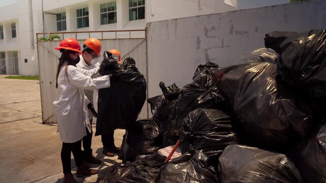Three workers, two women and one man, at a recycling plant move black trash bags for the recycling process. They are dressed in white lab coats and wear safety equipment, including hard hats and face