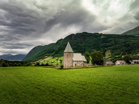 Aerial view of Hove Church surrounded by lush green fields and dramatic mountains under a cloudy sky in Vik i Sogn, Vestland, Norway.