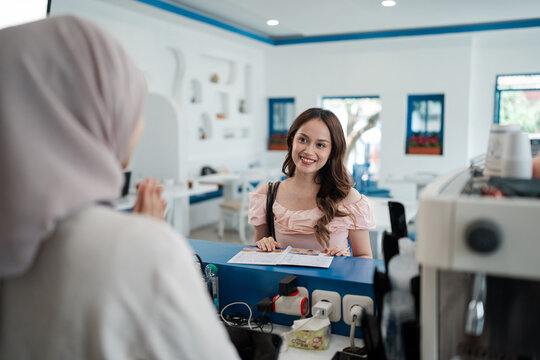 Smiling young woman chats with receptionist at clinic counter, holding brochure while arranging appointment in bright modern healthcare office with clean blue accents and soft daylight.